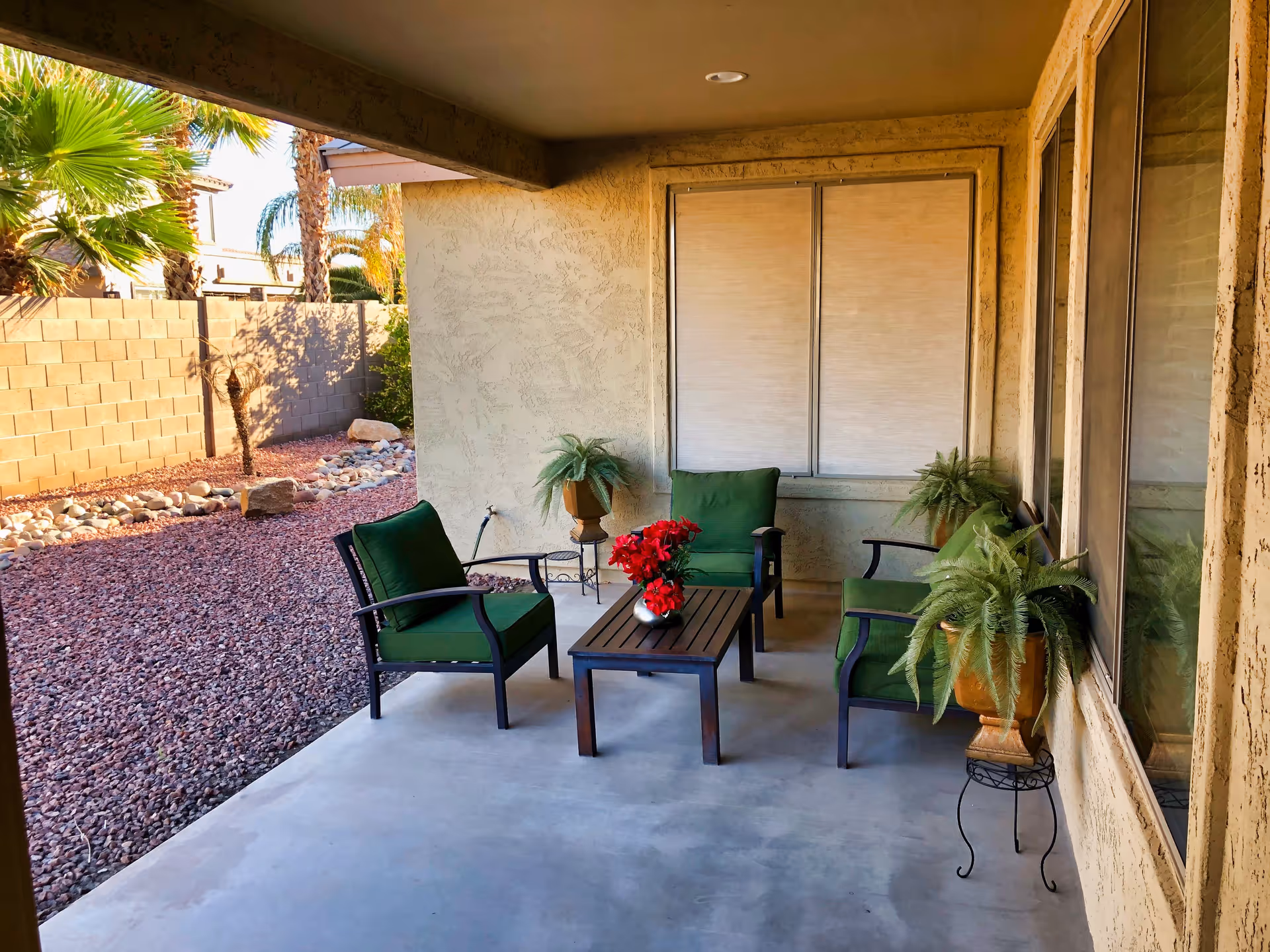 Covered outdoor patio area with four green cushioned chairs arranged around a small wooden coffee table with a red flower centerpiece. There are potted plants on stands near the chairs and a window with closed blinds on the wall. The patio overlooks a yard with red gravel, rocks, and palm trees, enclosed by a beige brick wall.