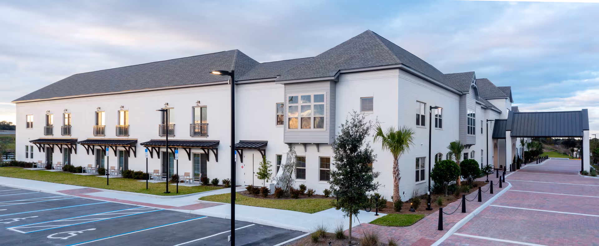 Exterior view of a large, two-story white building with multiple windows and small balconies. The building has a gray roof and is surrounded by landscaped greenery including trees and bushes. There is a paved parking area with marked handicap spaces and a covered entrance walkway on the right side. The sky is partly cloudy.