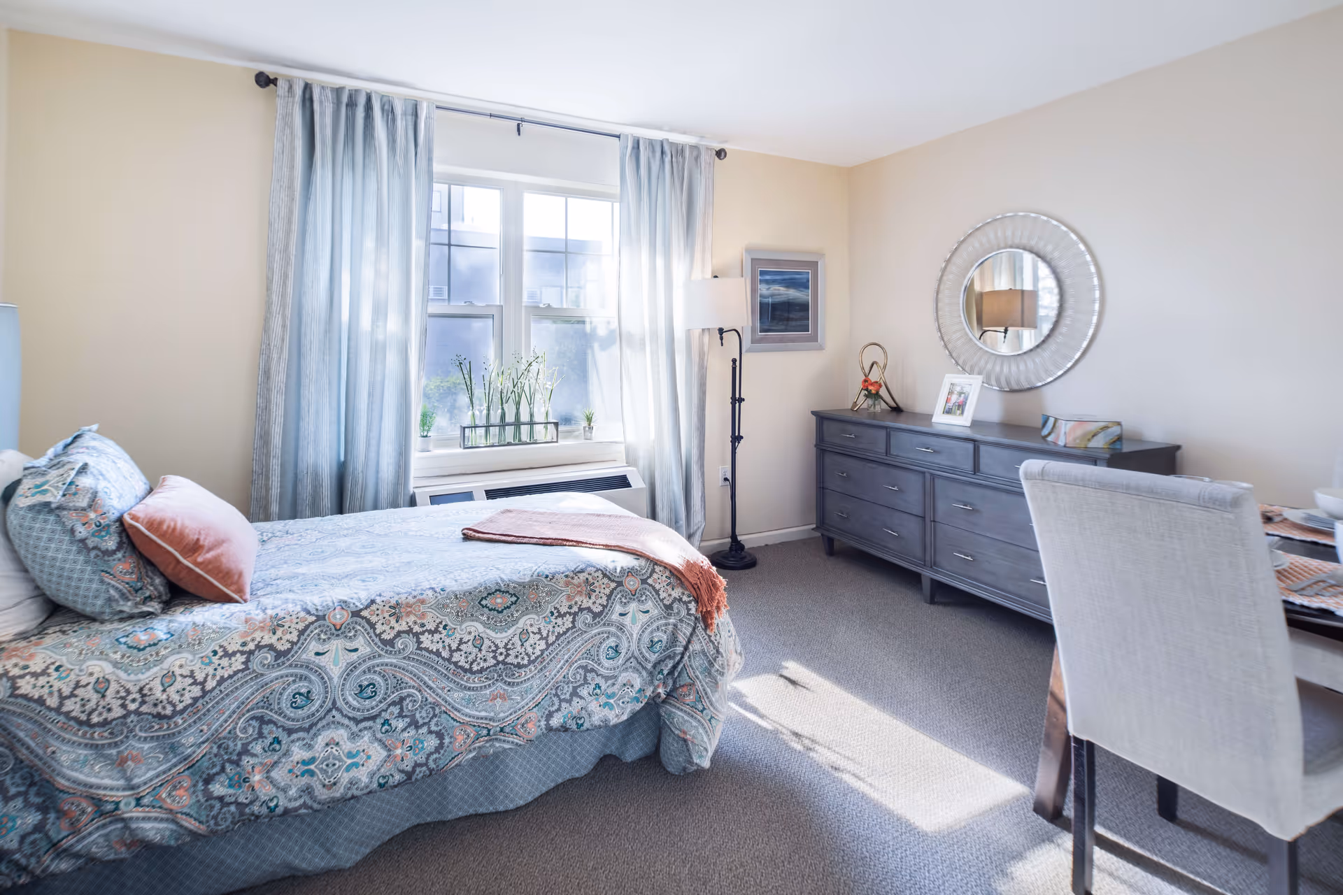 A bright bedroom with a bed covered in a patterned blue and coral bedspread, a window with light blue curtains letting in natural light, a gray dresser with a round mirror above it, a floor lamp, and a dining table with a beige upholstered chair.