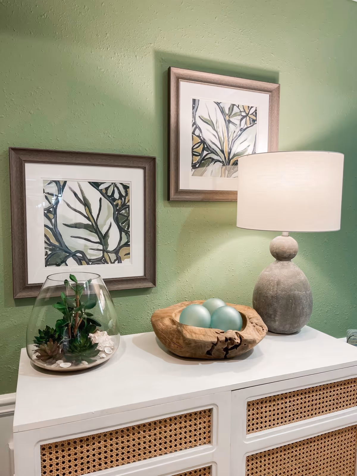 Console table against a green wall with two framed botanical prints, a lamp, a wooden bowl of decorative spheres, and a glass terrarium.