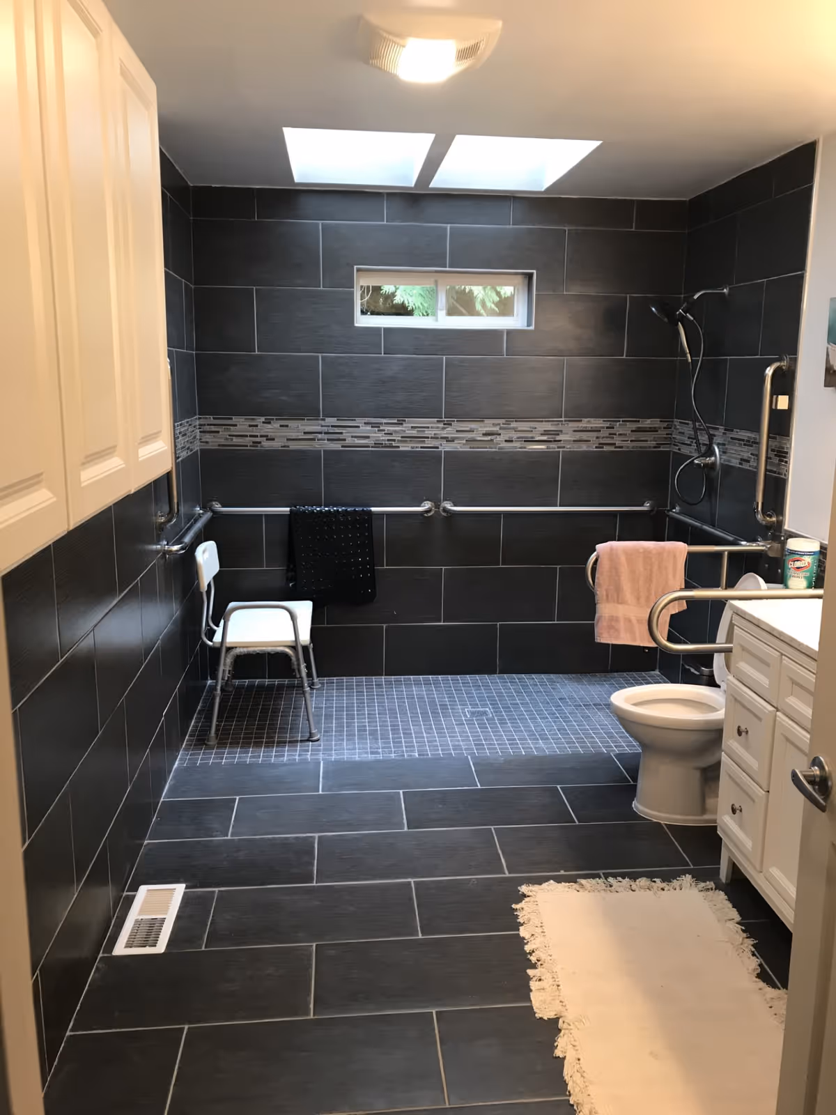 A modern bathroom with dark gray tiles on the walls and floor. The shower area has a small white chair, grab bars, and a handheld showerhead. There is a small window above the shower area and two skylights on the ceiling. A white toilet with grab bars and a white vanity with drawers are visible on the right side. A white rug is placed on the floor near the vanity.