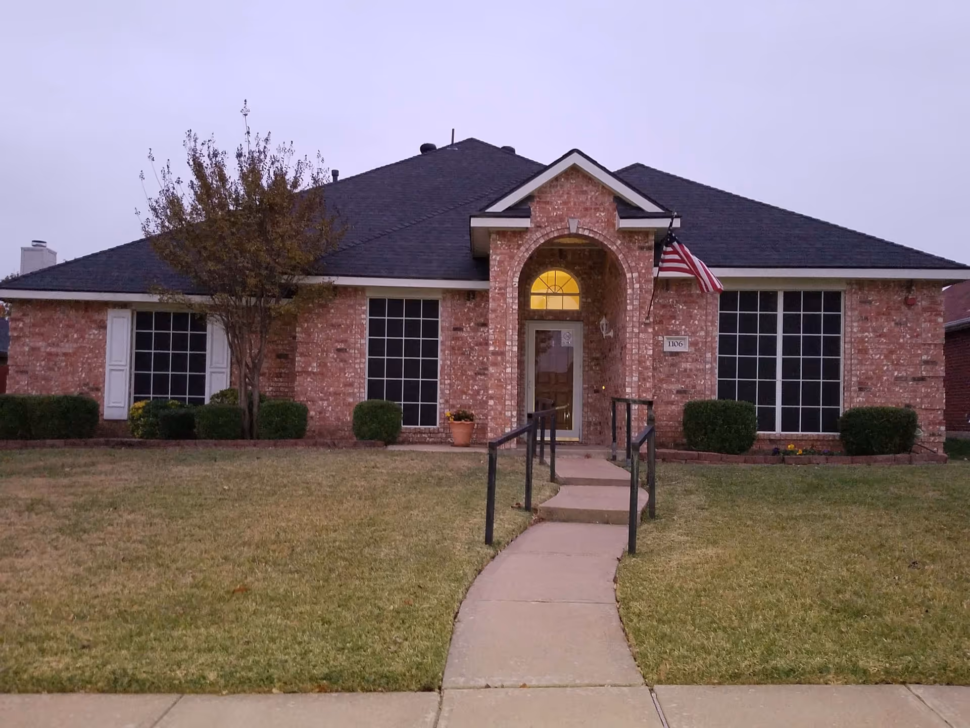 Front exterior view of a single-story brick building with a dark shingled roof, large windows, a central arched entrance with a glass door, an American flag mounted near the entrance, and a concrete walkway with handrails leading up to the door. The lawn is neatly trimmed with some bushes and a small tree near the building.