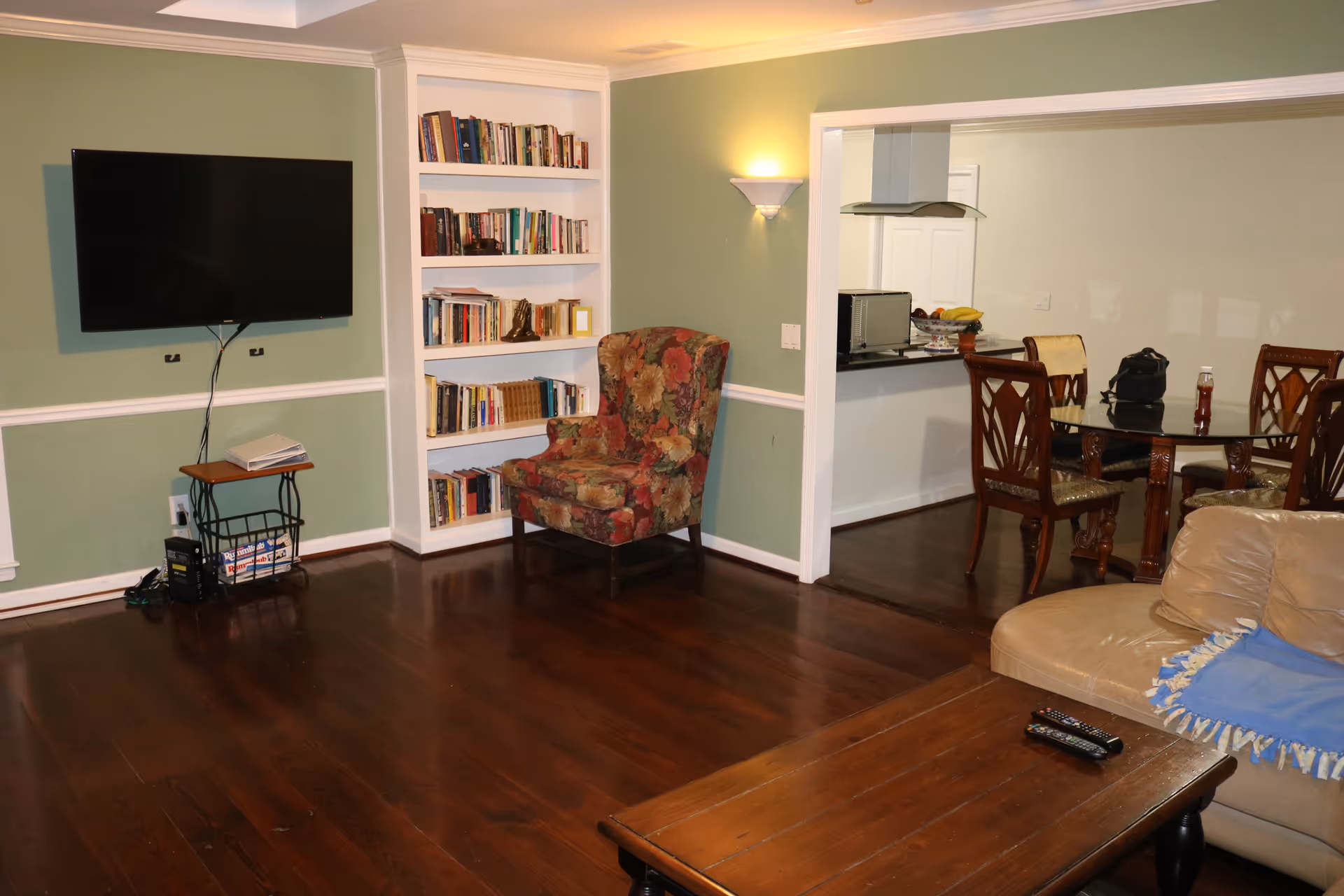 Interior view of a living room with green walls, a mounted flat-screen TV, a floral upholstered armchair, a built-in white bookshelf filled with books, a wooden coffee table with remote controls, and a beige leather sofa with a blue throw blanket. In the background, there is a dining area with a glass-top table and wooden chairs, and a kitchen counter with a microwave and a fruit bowl.