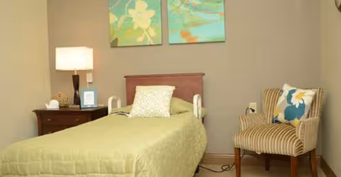 A single bed with a green bedspread and a patterned pillow in a room with beige walls. Next to the bed is a wooden nightstand with a lamp, a small decorative item, and a framed picture. On the wall above the bed are two colorful floral paintings. To the right of the bed is a striped armchair with a floral cushion.