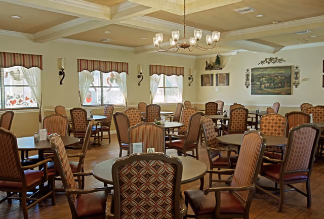 A spacious dining room with multiple round tables and cushioned chairs arranged neatly. The room has large windows with sheer white curtains and striped valances, allowing natural light to fill the space. The walls are decorated with framed artwork and sconces, and a chandelier hangs from the coffered ceiling. The floor is wooden, creating a warm and inviting atmosphere.