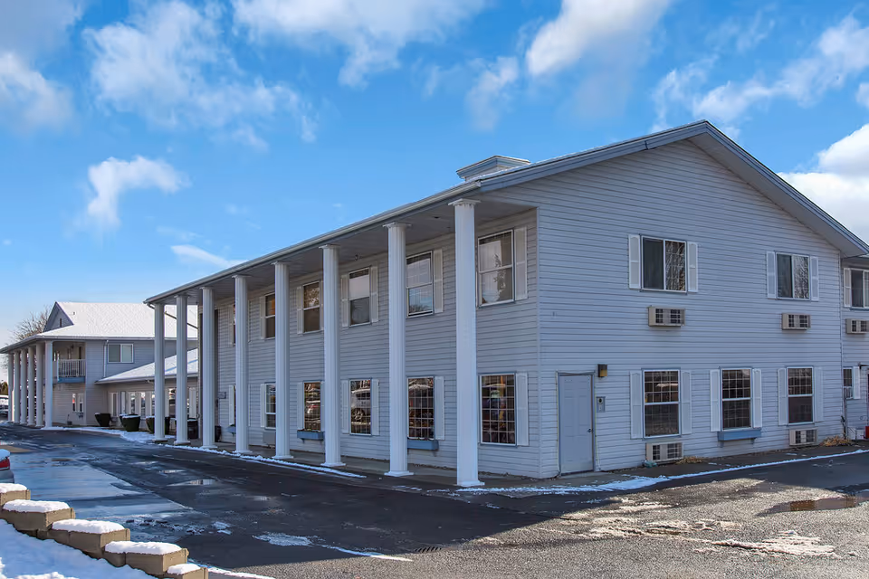 Two-story light-gray senior living building with white columns, many windows, and a parking area with patches of snow under a blue sky.