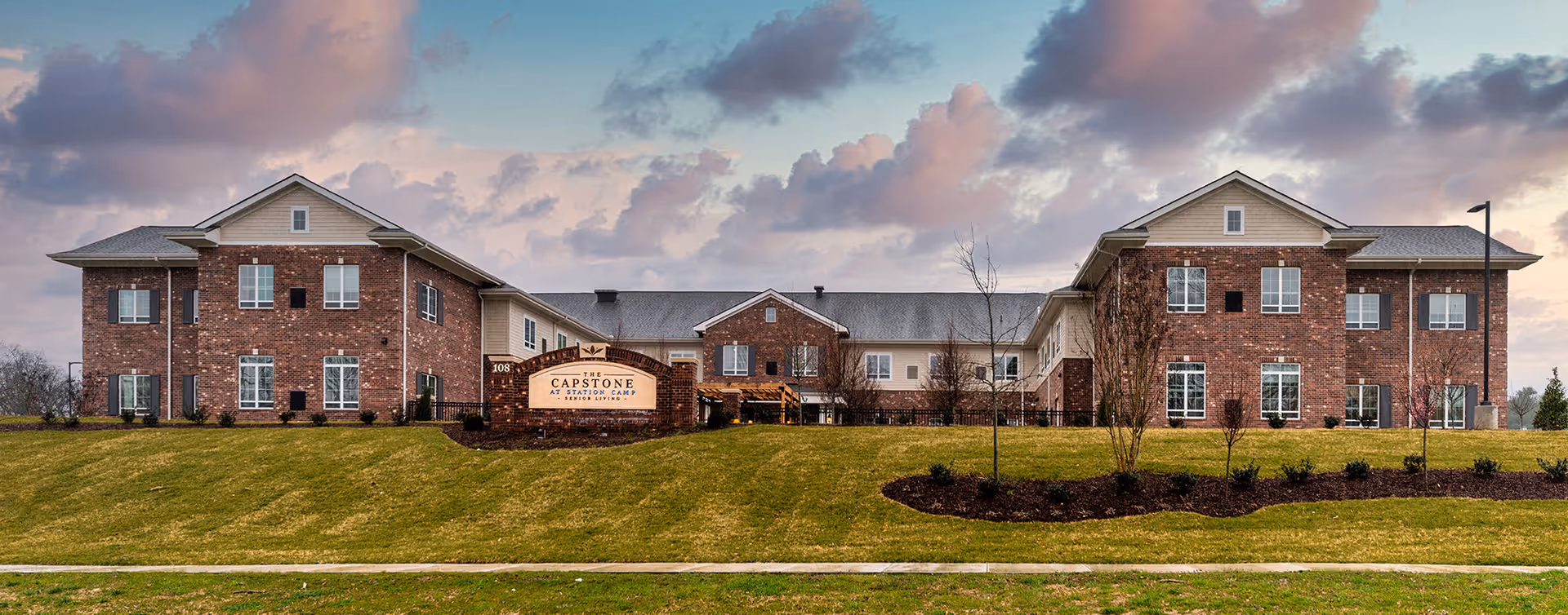 Exterior front view of The Capstone at Station Camp senior living facility, a two-story brick building with multiple windows, a landscaped lawn, and a sign in front displaying the facility's name.