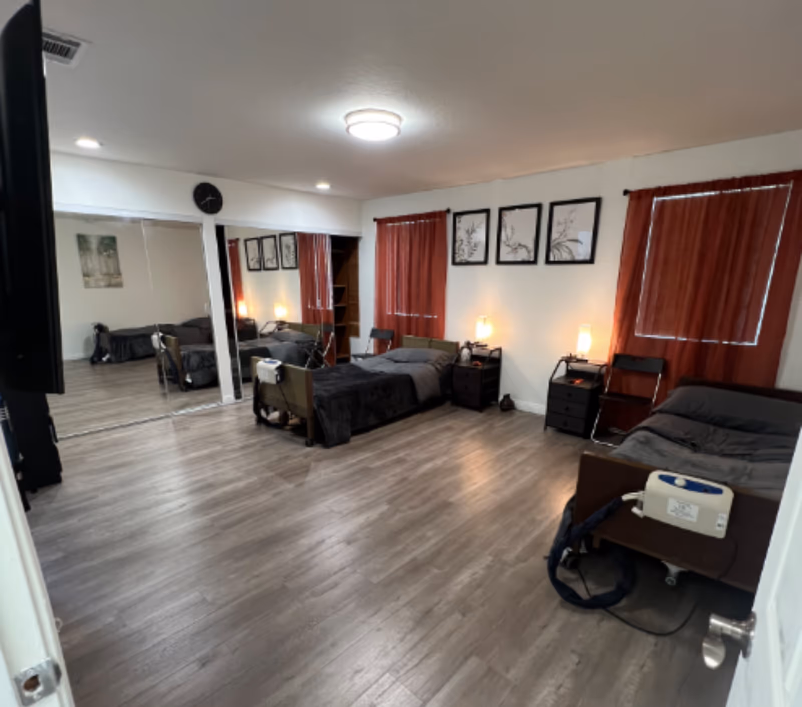 Large shared bedroom with several beds, nightstands, lamps, red curtains and mirrored closet on wood-look flooring.