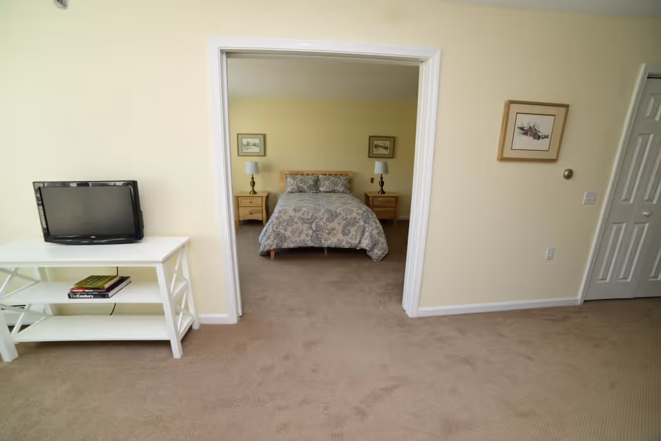 View of a bedroom through an open doorway from an adjacent room. The bedroom has a bed with a patterned bedspread, two nightstands with lamps, and framed pictures on the wall. In the foreground, there is a white TV stand with a flat-screen TV and some books on the lower shelf. The walls are painted light yellow and the floor is carpeted in beige.