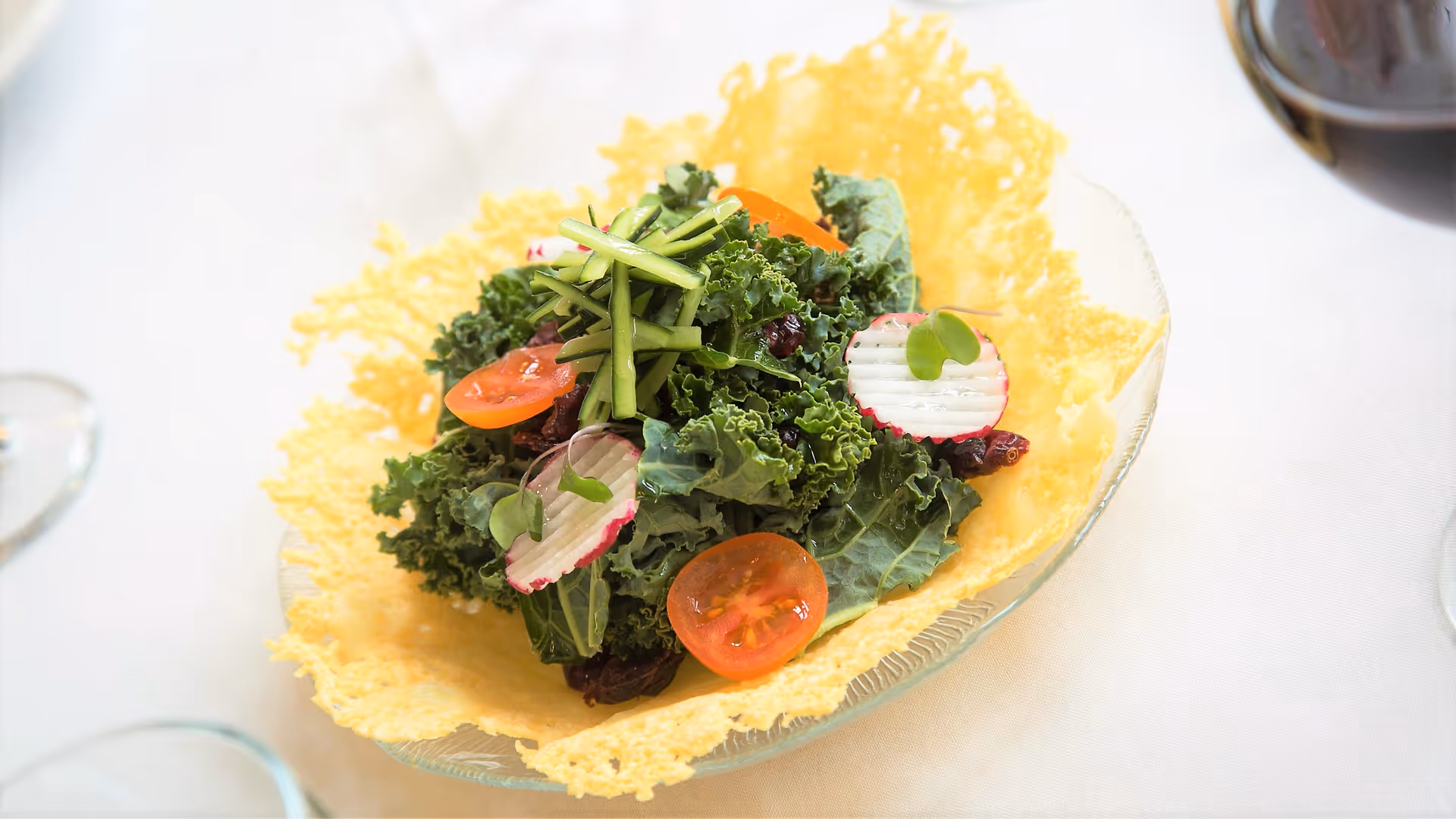 A fresh salad with kale, cherry tomatoes, radish slices, and thinly sliced green vegetables served in a crispy cheese bowl on a white tablecloth.