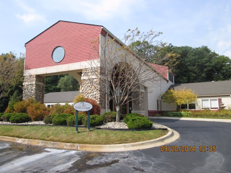 Exterior view of a building entrance with stone pillars and red siding. There is a circular window above the entrance and a 'Welcome' sign in front surrounded by bushes and trees. The driveway curves around the landscaped area.