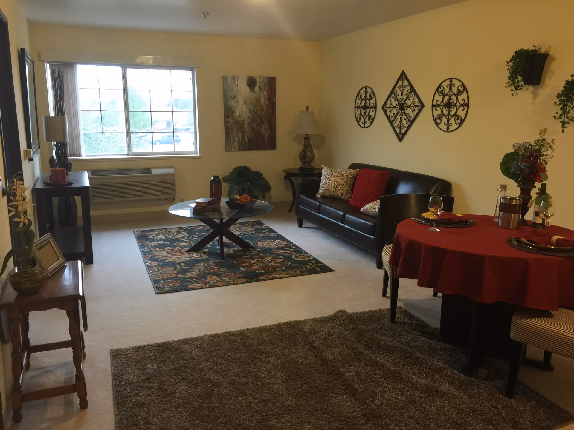 Bright living room with a leather sofa, decorative wall art, a glass coffee table on a patterned rug, and a small dining table set with a red tablecloth.