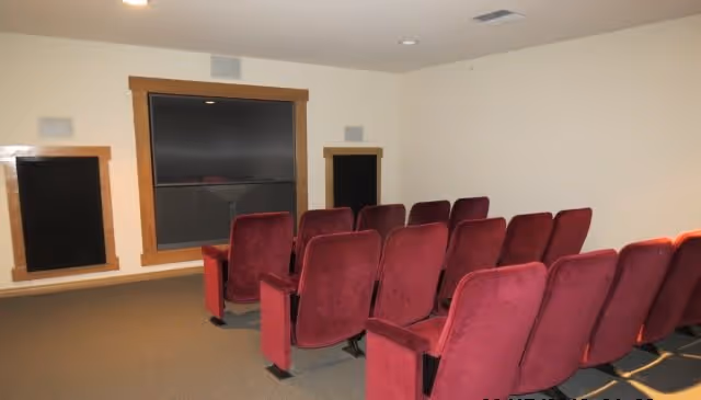Small screening room with rows of red upholstered theater seats facing a large wall-mounted screen and speakers.