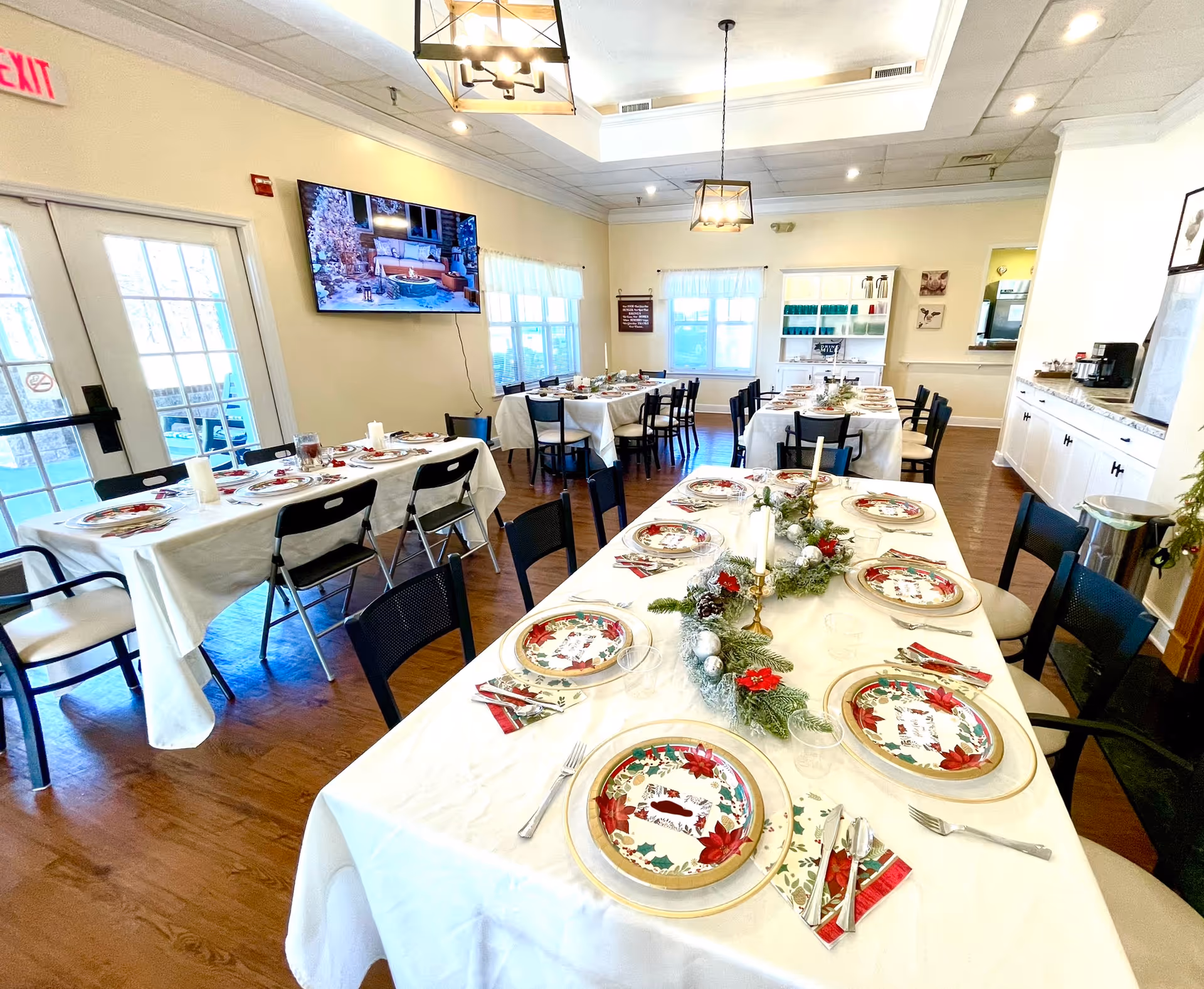 Dining room in an assisted living facility with long tables set for a holiday meal, decorated plates, candles and greenery.