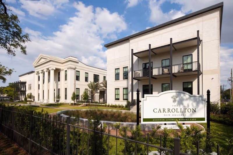 Front exterior of The Carrollton senior living facility showing its sign, landscaped courtyard, and buildings with classical columns and balconies.