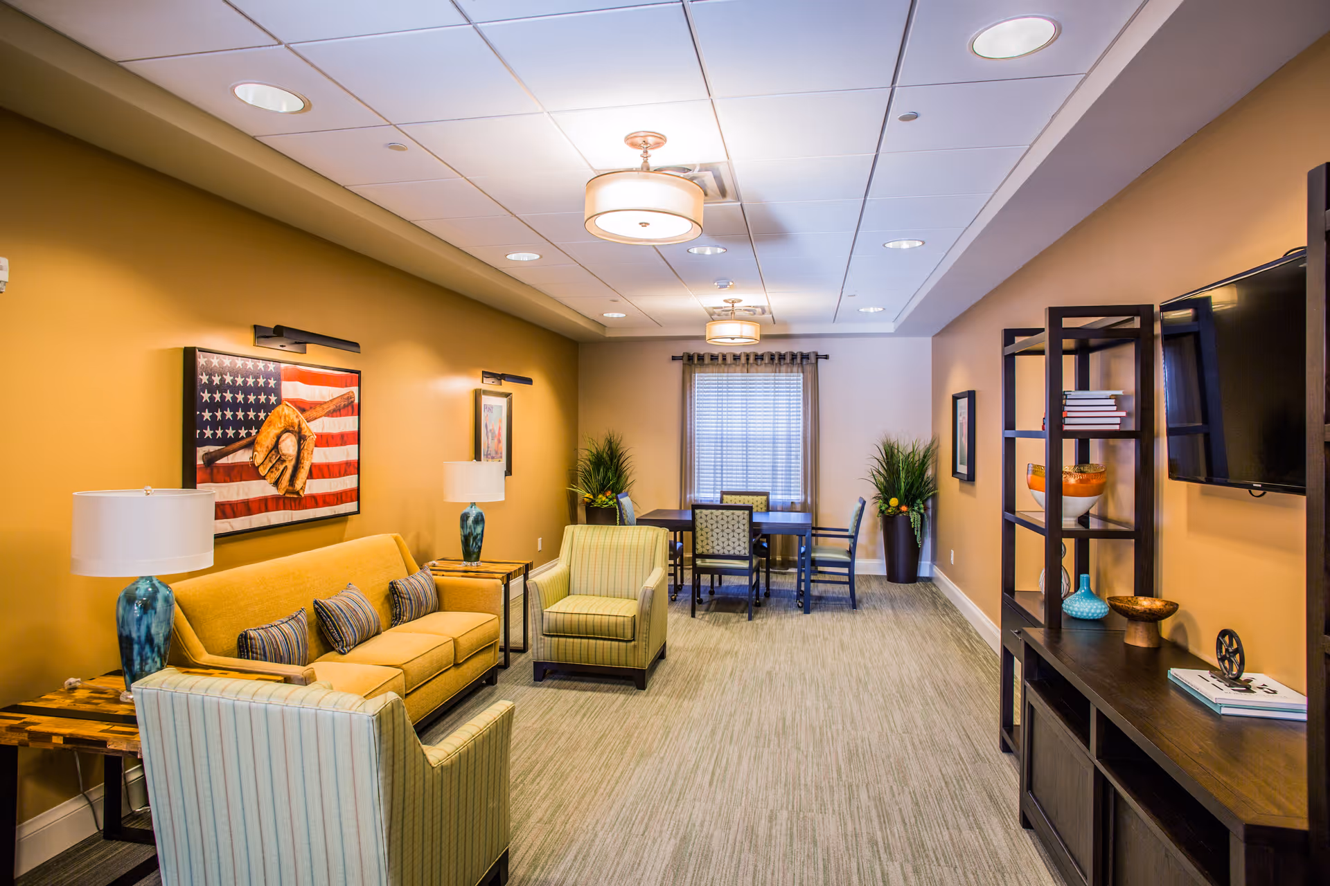 Warmly decorated lounge with a sofa and armchairs facing a wall-mounted TV, shelving, and a dining table near the window.