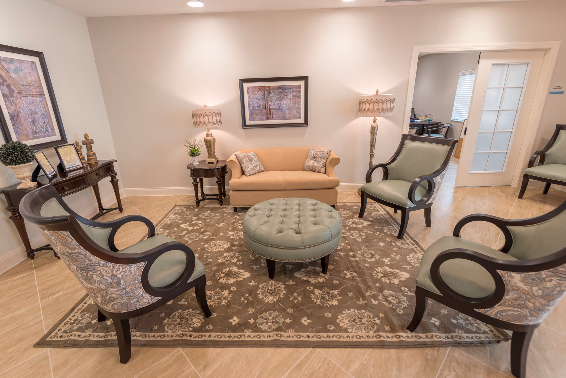 A cozy living room area with a beige loveseat, four green upholstered armchairs arranged around a round tufted ottoman on a floral patterned rug. The room has light-colored walls with framed artwork, two table lamps on side tables, and a console table with decorative items and picture frames. A doorway with a glass-paneled door leads to another room.
