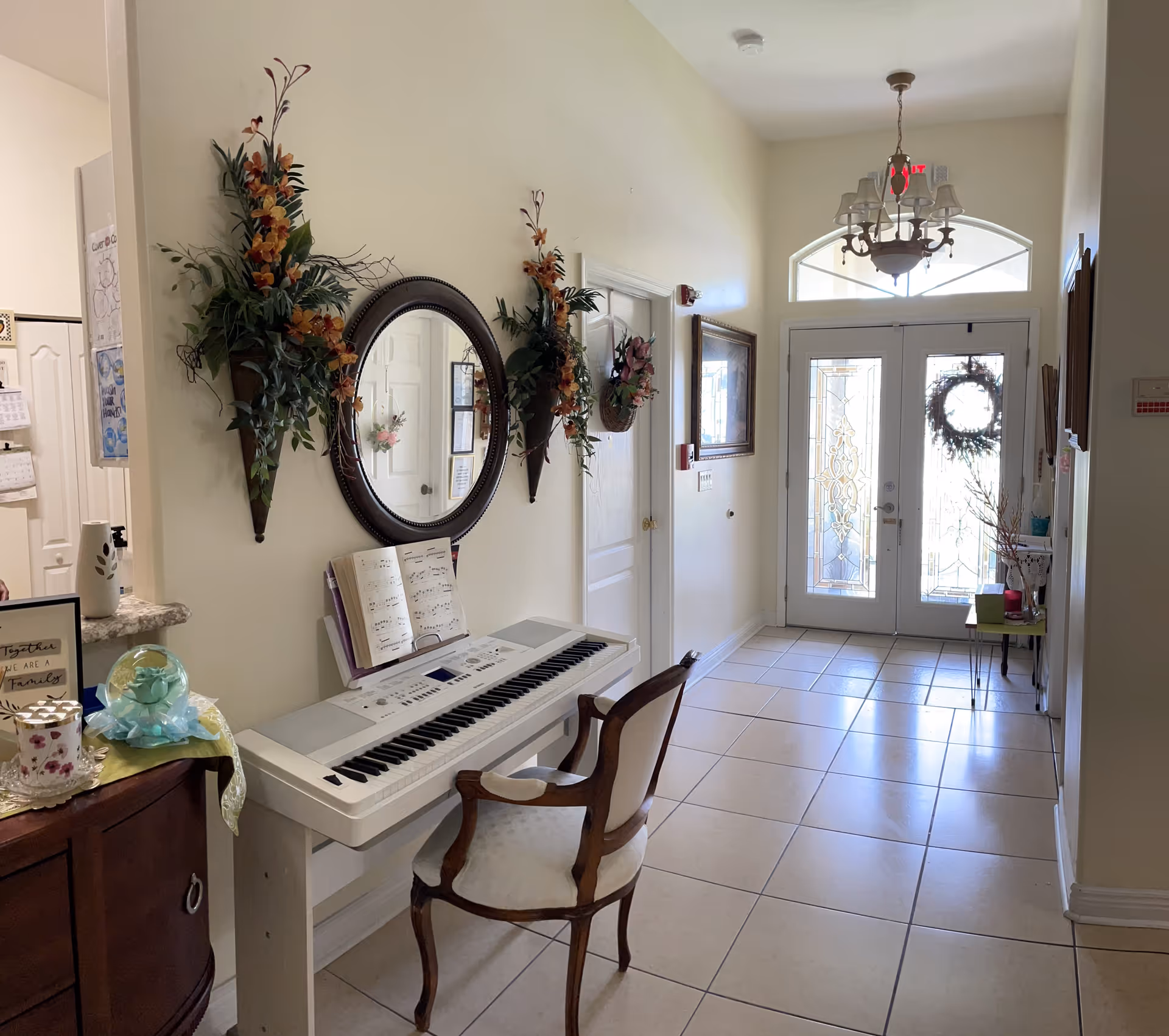 A bright hallway with tiled floor leading to a glass double door decorated with a wreath. On the left side, there is a white electronic keyboard with an open music book and a wooden chair with a cushioned seat. Above the keyboard, a round mirror is flanked by two wall-mounted floral arrangements. A chandelier hangs from the ceiling near the door. Various decorative items and framed pictures are visible along the walls and surfaces.