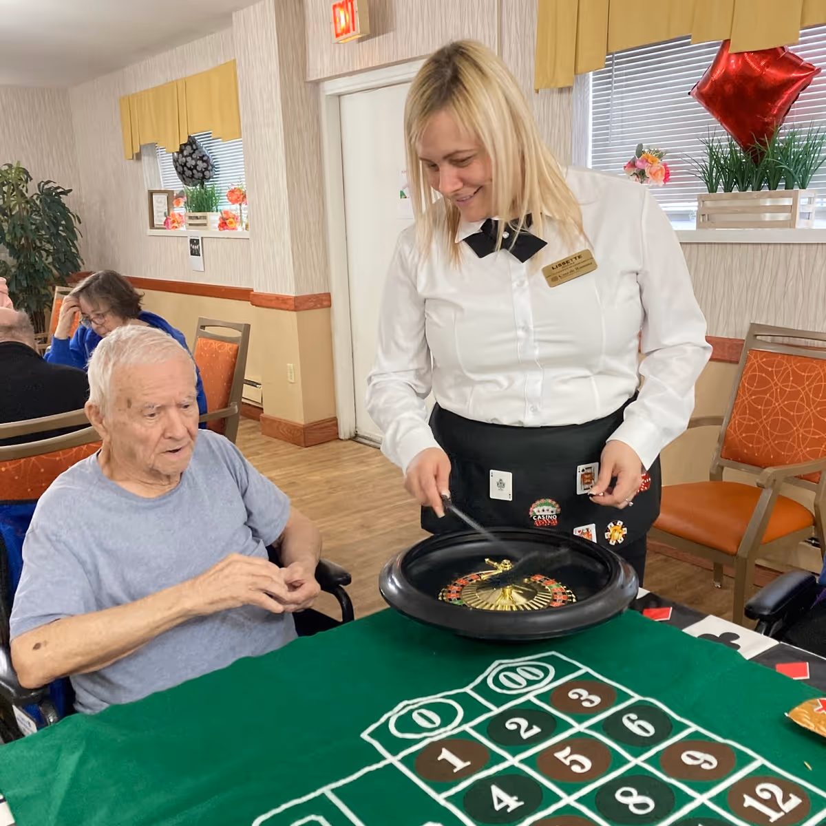 An elderly man in a wheelchair playing a roulette game at a green felt table with numbered betting areas, assisted by a smiling woman in a white shirt and black bow tie who is spinning the roulette wheel. The setting is a room with orange chairs, a window with yellow valances, and some plants and decorations in the background.