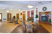 Bright communal dining and lounge area with round tables and chairs, a fireplace with a clock above, chandeliers, and wood flooring.