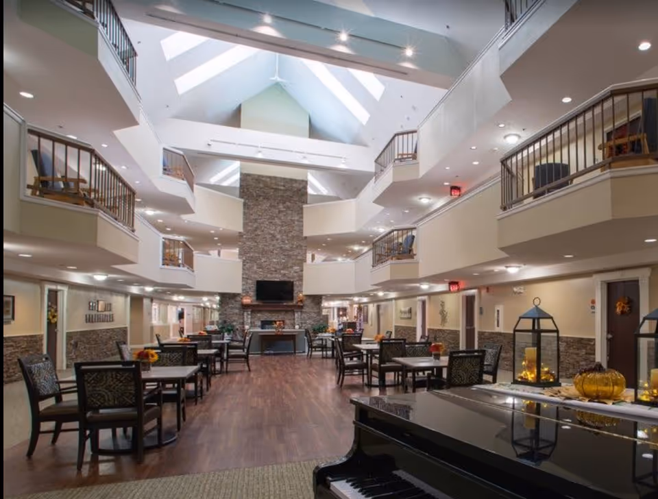 Spacious and well-lit common area in a senior living facility with multiple tables and chairs arranged for socializing or dining. The room features a high vaulted ceiling with skylights, a large stone fireplace with a mounted TV above it, and balconies overlooking the area. A black grand piano is visible in the foreground, and decorative lanterns and autumn-themed decorations are placed on tables.