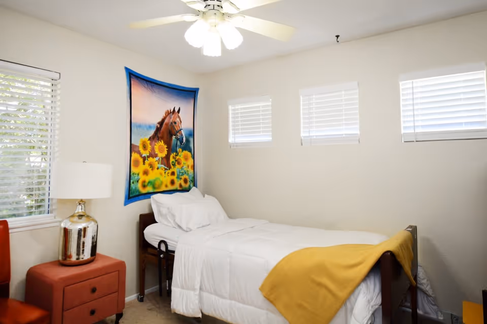 A small bedroom with a single bed covered in white bedding and a yellow blanket at the foot. Above the bed, there is a wall hanging featuring a horse and sunflowers. The room has three small windows with white blinds and a ceiling fan with lights. A red nightstand with a silver lamp is next to the bed.