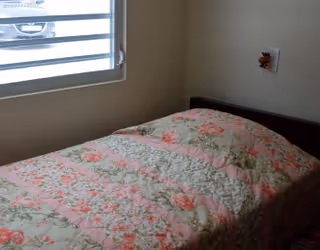 A small bedroom with a single bed covered in a floral patterned quilt in shades of pink and green. There is a window with horizontal blinds partially open, letting in natural light. A small stuffed toy is hanging on the wall near the head of the bed.