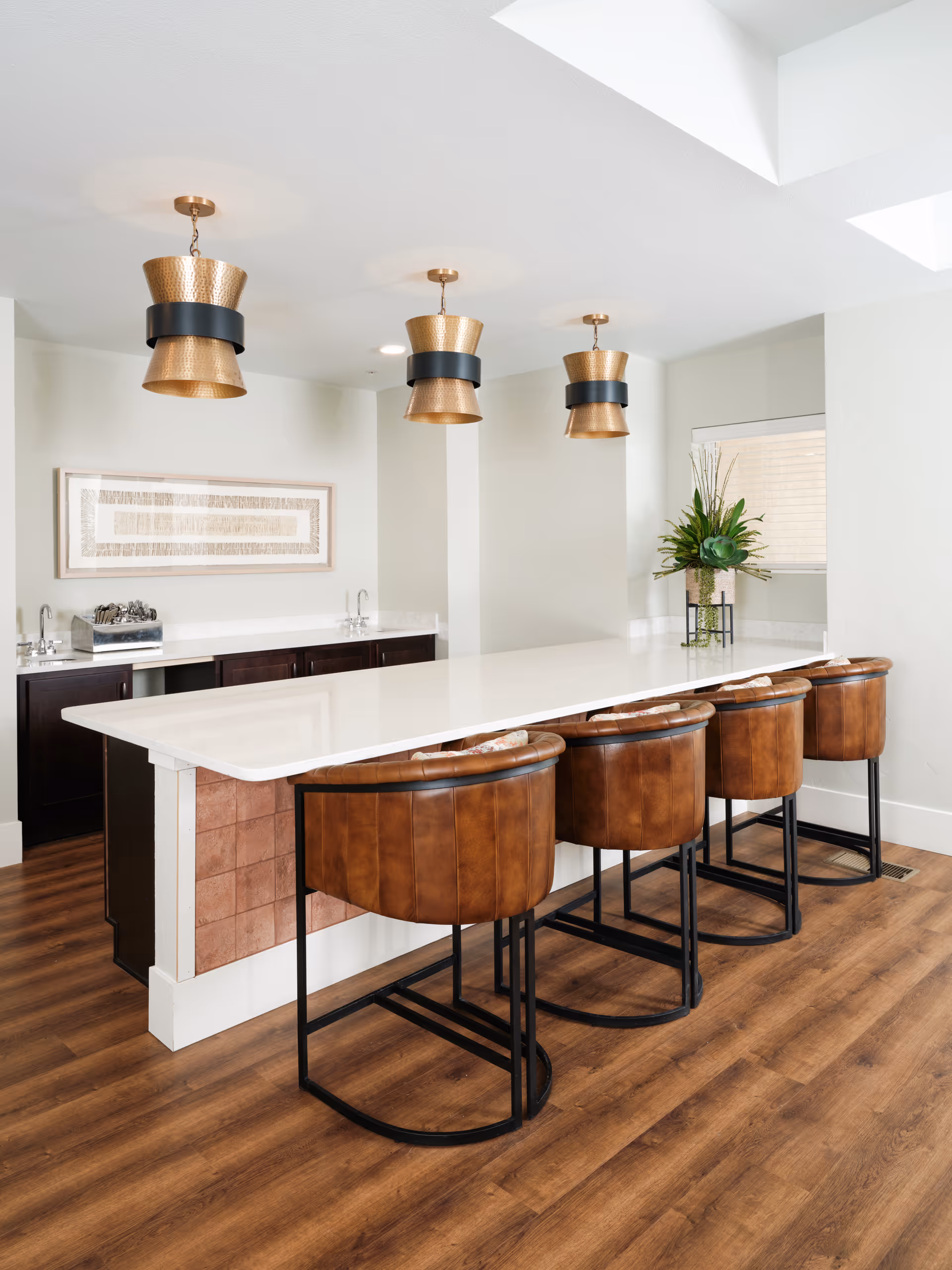 Bright communal kitchen/bar area with a long white countertop, four brown leather bar stools, pendant lights, and wood flooring.