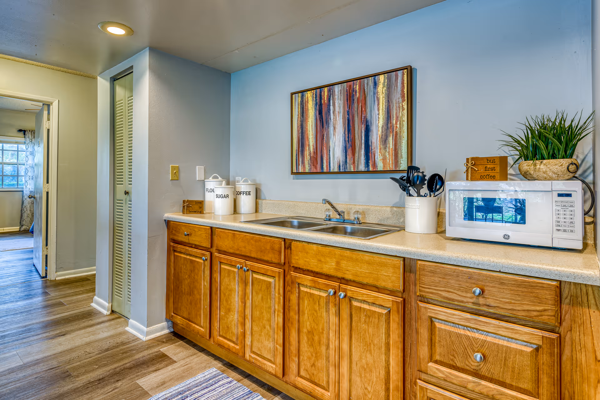 Kitchenette with wooden cabinets, a double sink, microwave, utensil holder and decorative artwork above the countertop.