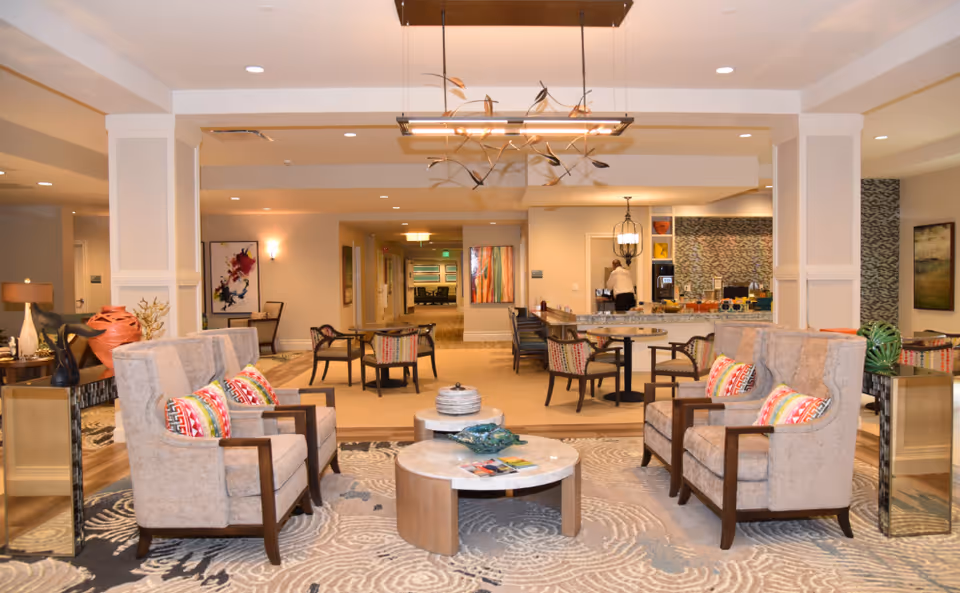 Spacious, well-lit senior living common area with paired armchairs around coffee tables and a dining/serving area in the background.