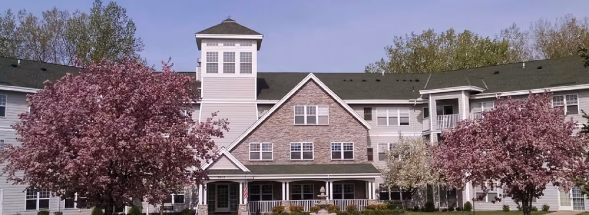 Front exterior view of Cardinal Pointe of Maplewood, a multi-story senior living facility with a central tower, stone and siding facade, surrounded by blooming pink and white trees under a clear blue sky.