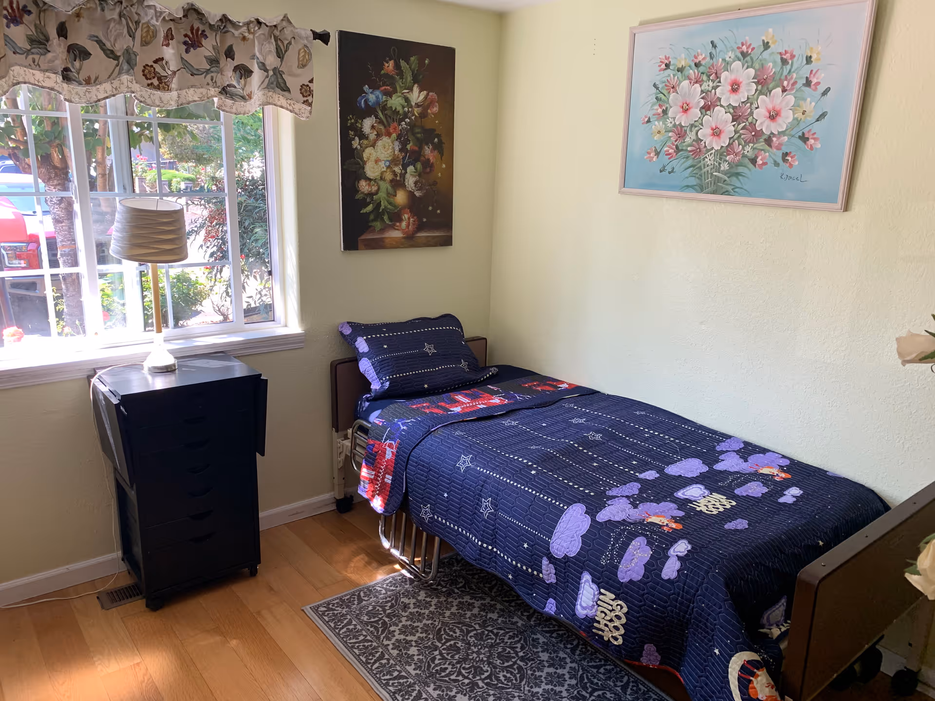 A small bedroom with a single bed covered in a dark blue quilt with purple and red patterns. There is a black bedside table with a lamp next to a window with floral curtains. Two floral paintings hang on the light yellow walls. The floor is wooden with a small patterned rug beside the bed.