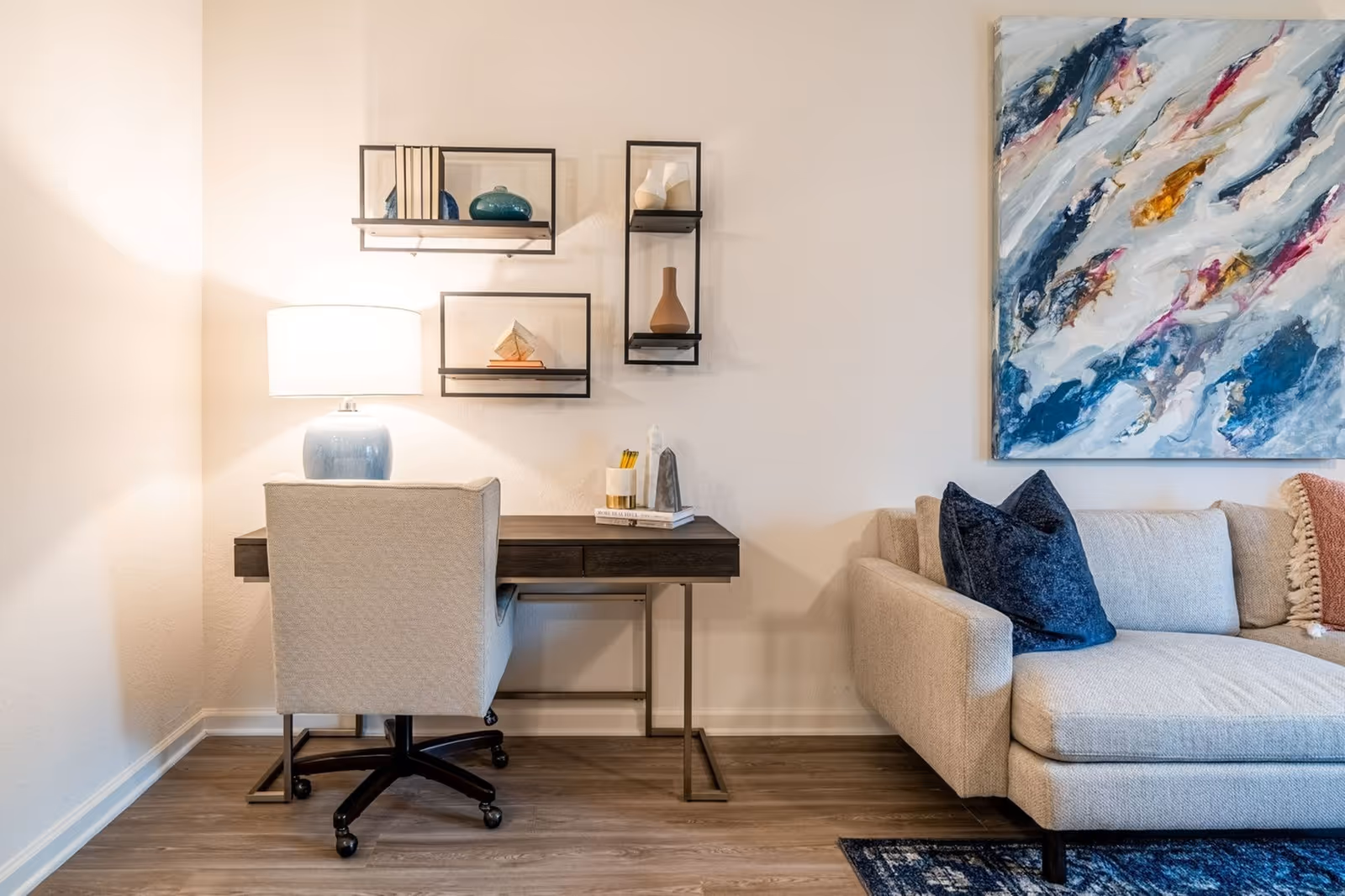 A cozy interior corner featuring a beige upholstered office chair on wheels in front of a dark wooden desk with a blue ceramic lamp, books, and decorative items. Above the desk are three black-framed wall shelves holding books and vases. To the right is a beige sofa with dark blue and beige cushions and a colorful abstract painting on the wall behind it. The floor is wooden with a blue patterned rug partially visible.