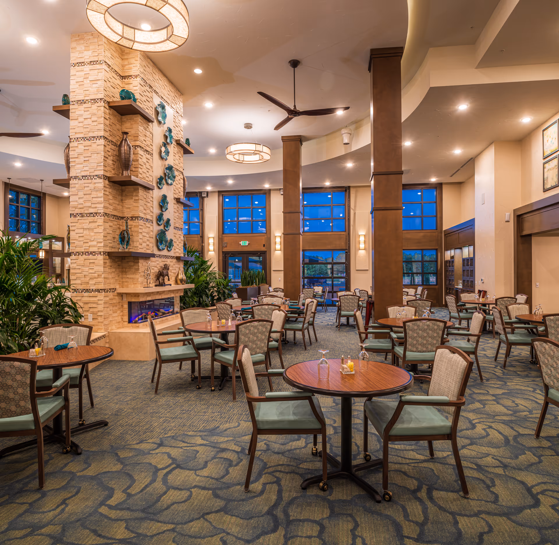 Spacious dining room with multiple round wooden tables and cushioned chairs arranged neatly. The room features a large stone fireplace with decorative shelves and blue ceramic wall art. Large windows with wooden frames let in natural light, and the ceiling has modern light fixtures and a ceiling fan. The carpet has a blue and beige pattern, and there are green plants placed around the room.