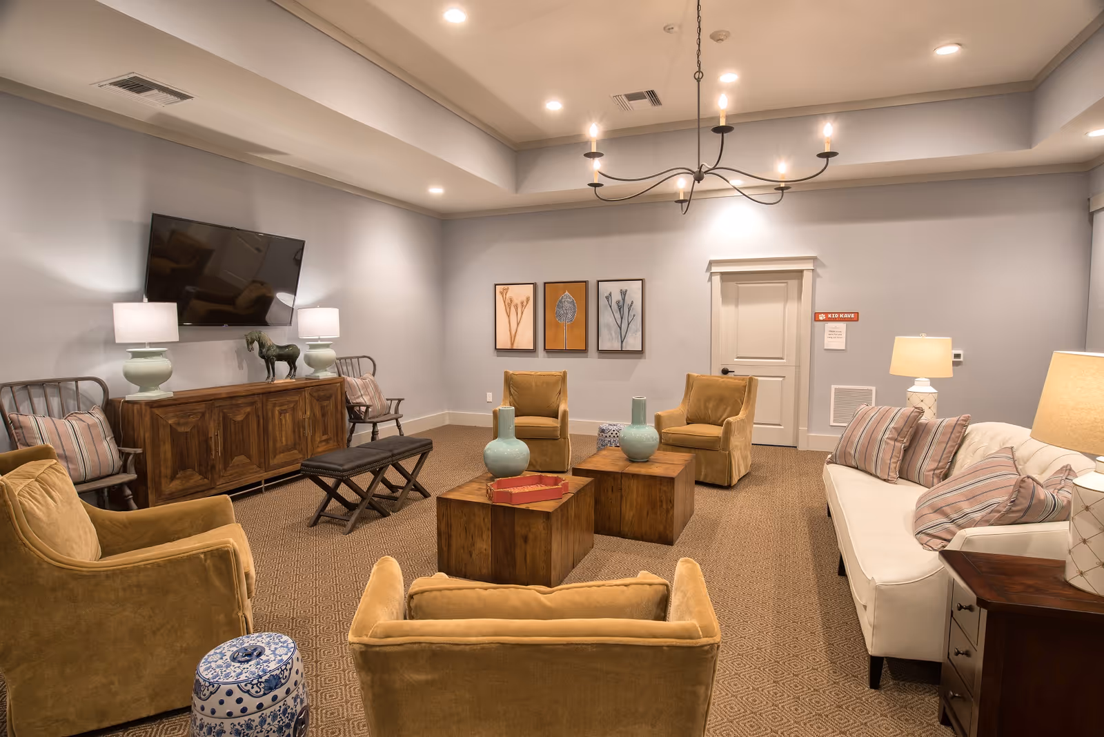 A cozy living room with beige and mustard yellow armchairs, a white sofa with striped pillows, wooden coffee tables with decorative vases, a wooden sideboard with two lamps and a horse figurine, a wall-mounted flat-screen TV, and framed botanical artwork on the wall. The room has soft lighting from ceiling fixtures and table lamps.