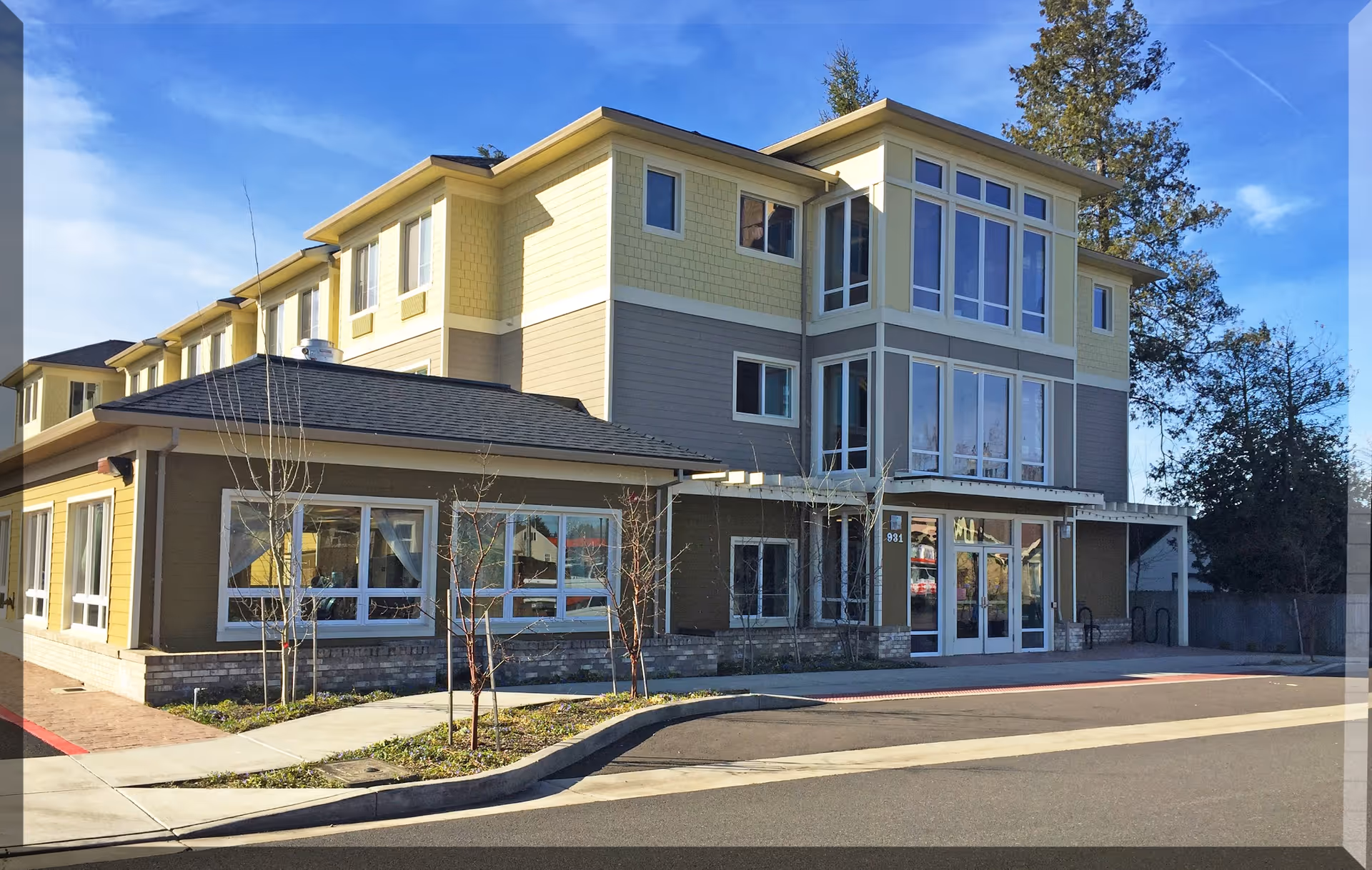 Exterior view of a modern three-story senior living facility building with large windows, a main entrance with glass doors, and a small landscaped area with young trees in front. The sky is clear and blue.