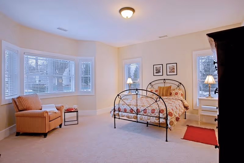 A bright bedroom with beige walls and carpet, featuring a metal-framed bed with a patterned quilt and pillows. There are two windows behind the bed with white blinds, each flanked by a small white nightstand with a lamp. To the left, there is a striped armchair with a white towel or blanket draped over one arm and a small side table with a cup and book. A red rug is placed on the floor near the bed, and a dark wooden dresser is partially visible on the right side.