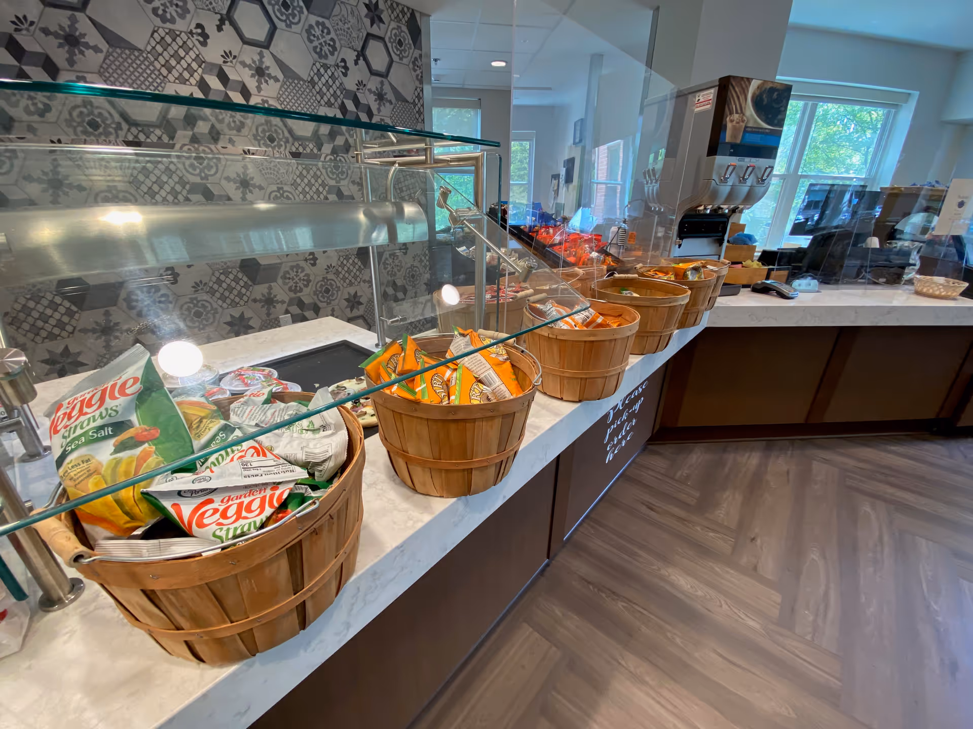 A snack bar area with multiple wooden baskets filled with various packaged snacks such as veggie straws and chips. The counter has a glass sneeze guard and a patterned tile backsplash. In the background, there are windows letting in natural light and a beverage dispenser.