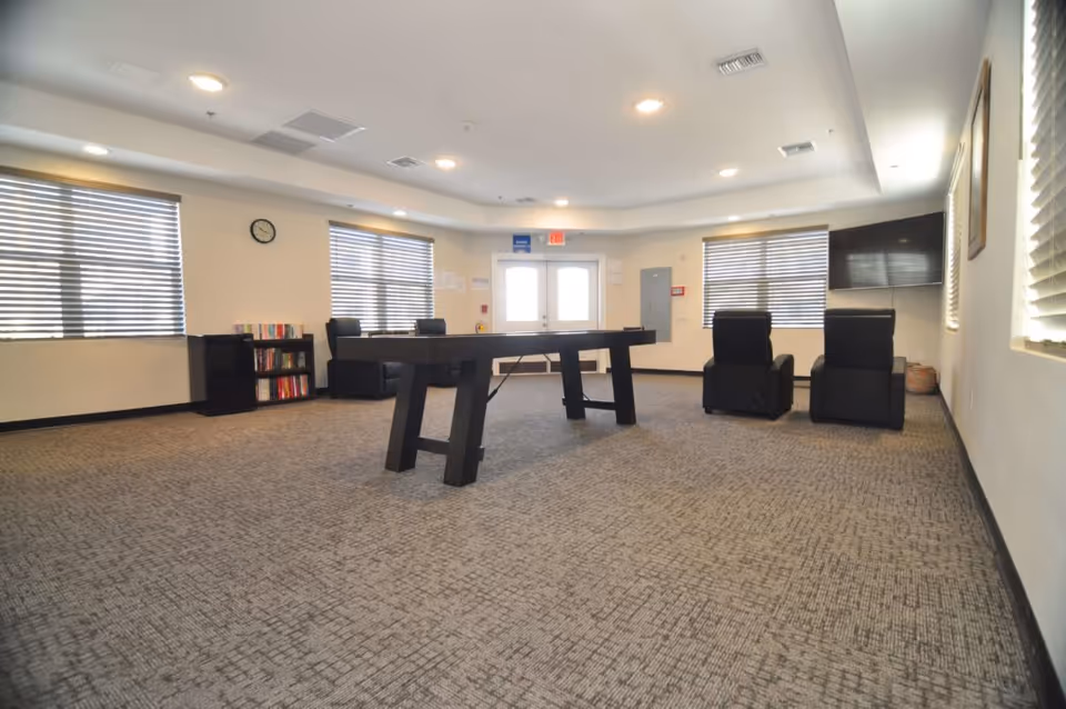 A spacious room with carpeted floor featuring a black foosball table in the center, two black armchairs facing a wall-mounted TV, bookshelves with books, multiple windows with blinds, and a clock on the wall above the bookshelves. The room has recessed lighting and a double door exit.