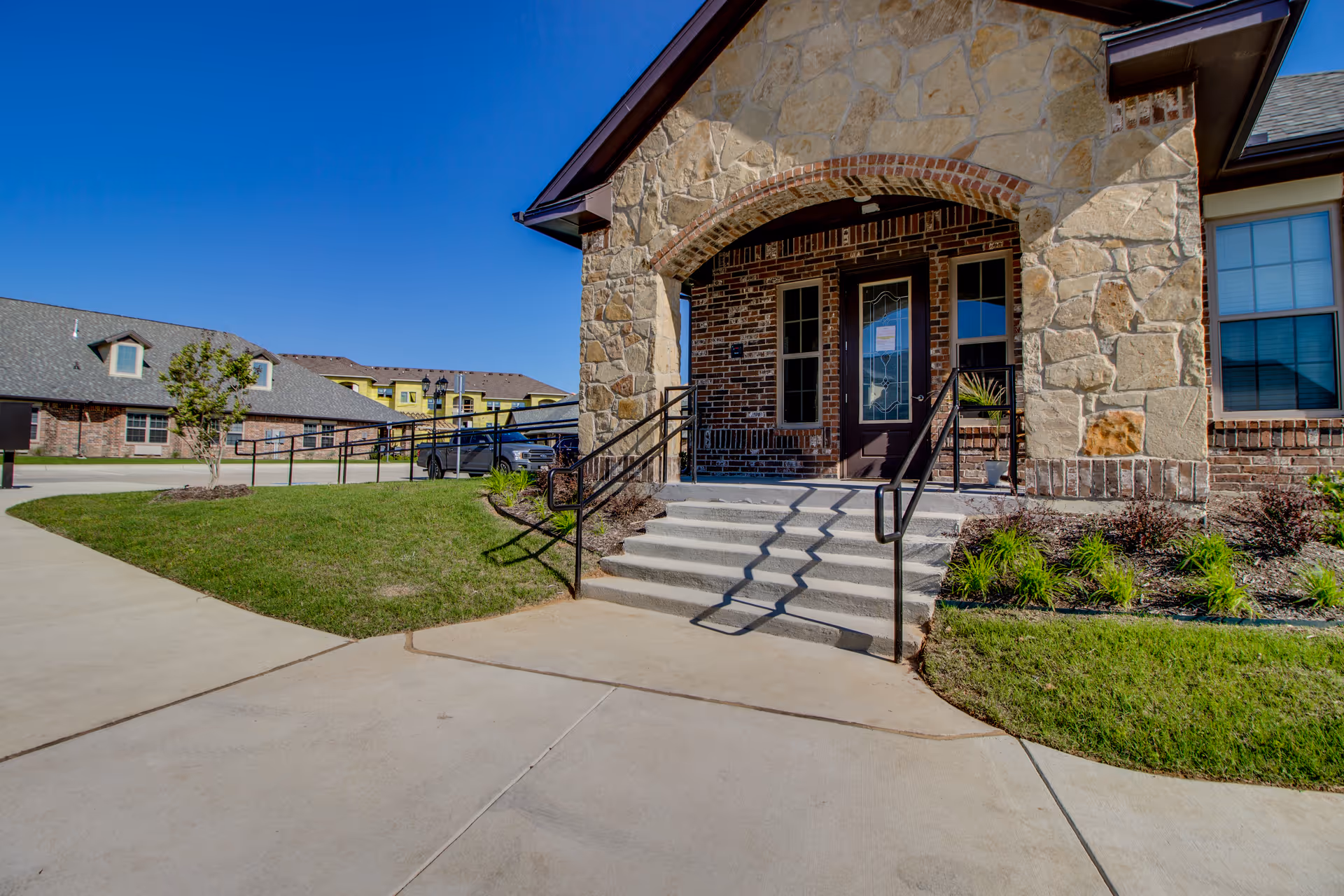 Entrance of a building with stone and brick exterior, concrete steps with black handrails leading to a glass door, surrounded by green grass and landscaping under a clear blue sky.