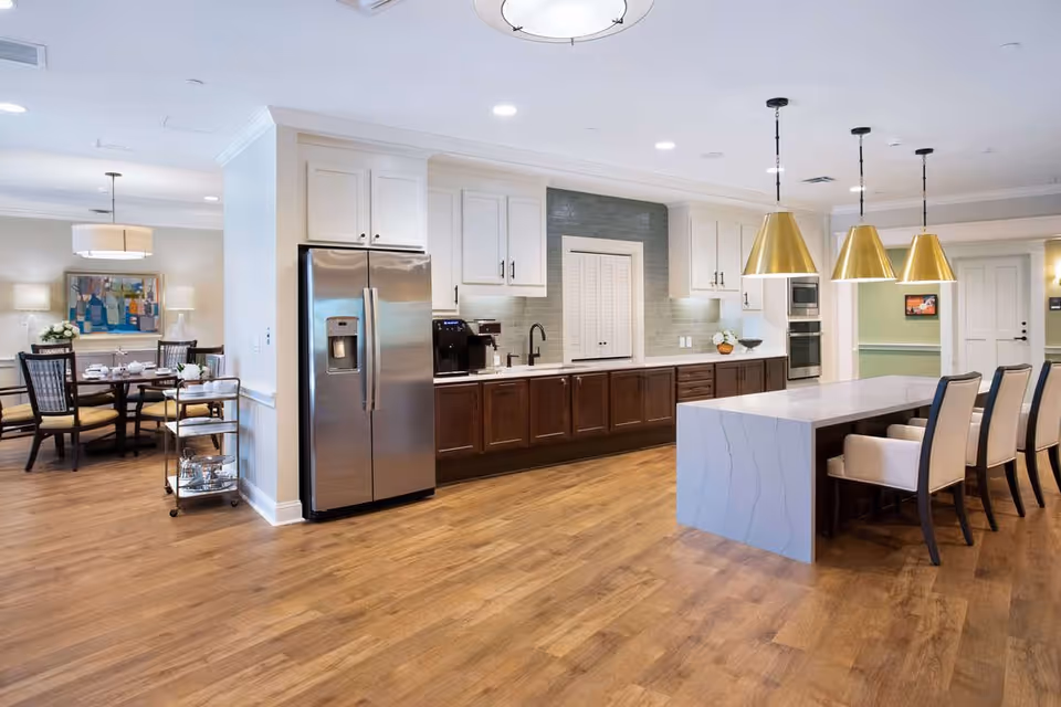A modern kitchen and dining area in a senior living facility with wooden flooring, white and dark brown cabinetry, a stainless steel refrigerator, a large kitchen island with white marble countertop, and four beige cushioned chairs. The dining area has a round table with chairs and is decorated with a painting and lamps.