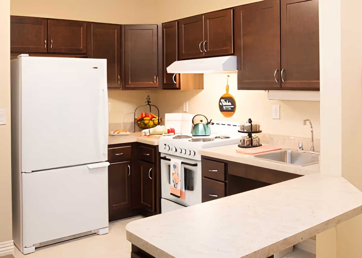 A kitchen with dark brown cabinets, a white refrigerator, a white stove with a teal kettle on it, a countertop with a two-tiered tray, a sink, and a fruit basket in the corner.