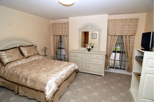A bedroom with a bed covered in a beige floral comforter and matching pillows. There are two windows with beige floral curtains on either side of a white dresser with a mirror and a flower arrangement on top. A small TV sits on a white shelving unit to the right. The room has beige walls and carpeted floor.