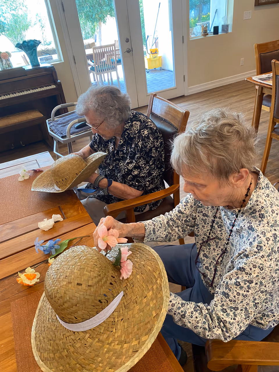 Two elderly women sitting at a wooden table indoors, decorating large straw hats with artificial flowers. One woman is placing pink flowers on her hat, while the other is holding her hat and looking down. The room has wooden flooring, a piano, chairs, and large glass doors leading outside.