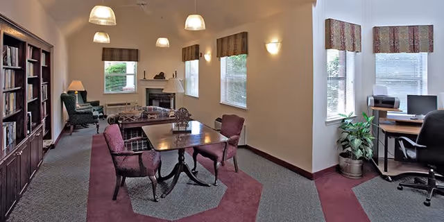 A cozy common area with a table and four chairs in the center, a fireplace with a mantel, bookshelves filled with books along one wall, and multiple windows with patterned valances letting in natural light. There is also a small desk with a computer and office chair near the windows, and a potted plant beside it.