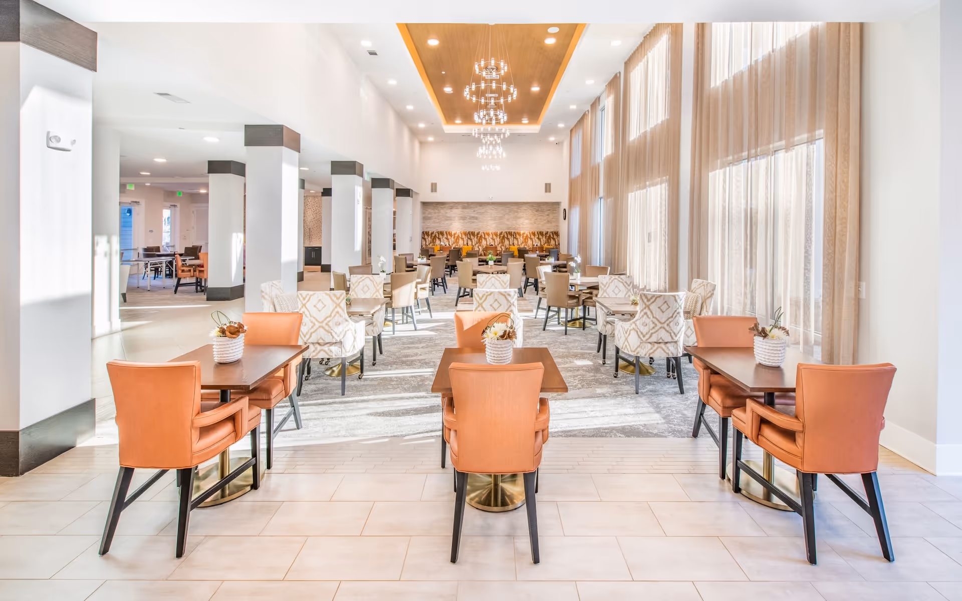 A bright and spacious dining room with multiple tables and chairs arranged neatly. The room features large windows with sheer beige curtains allowing natural light to fill the space. The ceiling has modern chandeliers and a wooden inset design. The tables are decorated with small flower arrangements.
