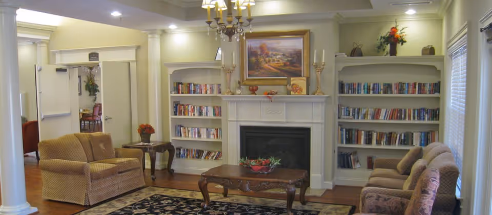 A cozy living room with two beige upholstered sofas facing each other across a wooden coffee table with a decorative centerpiece. The room features a white fireplace with a landscape painting above it, flanked by built-in bookshelves filled with books. There are decorative candlesticks on the mantel and a small side table with a flower arrangement. The room has hardwood floors and white columns near the entrance.