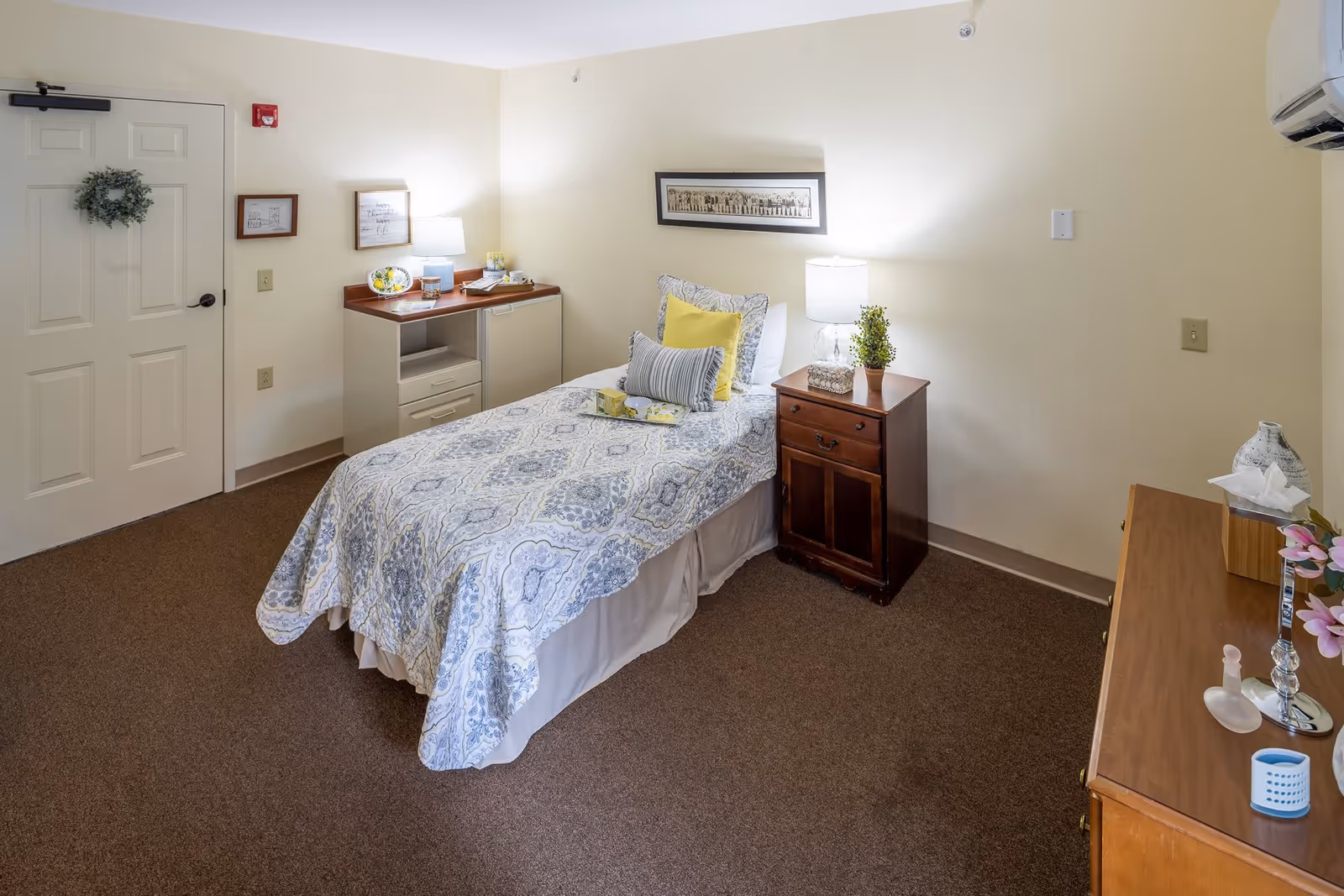 A neatly arranged bedroom in a senior living facility with a single bed covered in a patterned blue and white bedspread, accented with yellow and gray pillows. There is a wooden nightstand with a lamp and a small plant next to the bed. A dresser with decorative items and a tissue box is visible on the right side. The room has beige walls, brown carpet, and a white door with a small wreath hanging on it. A framed picture hangs above the bed, and a small cabinet with a lamp and decorative items is positioned against the wall near the door.