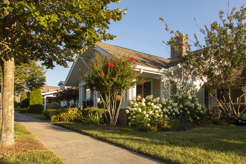 Exterior view of a single-story residential building with a porch, surrounded by well-maintained landscaping including trees, bushes, and flowering plants under a clear blue sky.