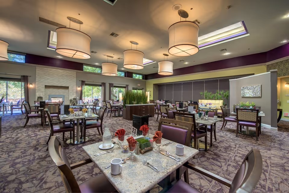 Spacious dining room with multiple tables set with cups, glasses, and red napkins. The room features large hanging light fixtures, carpeted floor with a patterned design, large windows with curtains, and a fireplace. There are plants and decorative elements around the room, creating a warm and inviting atmosphere.